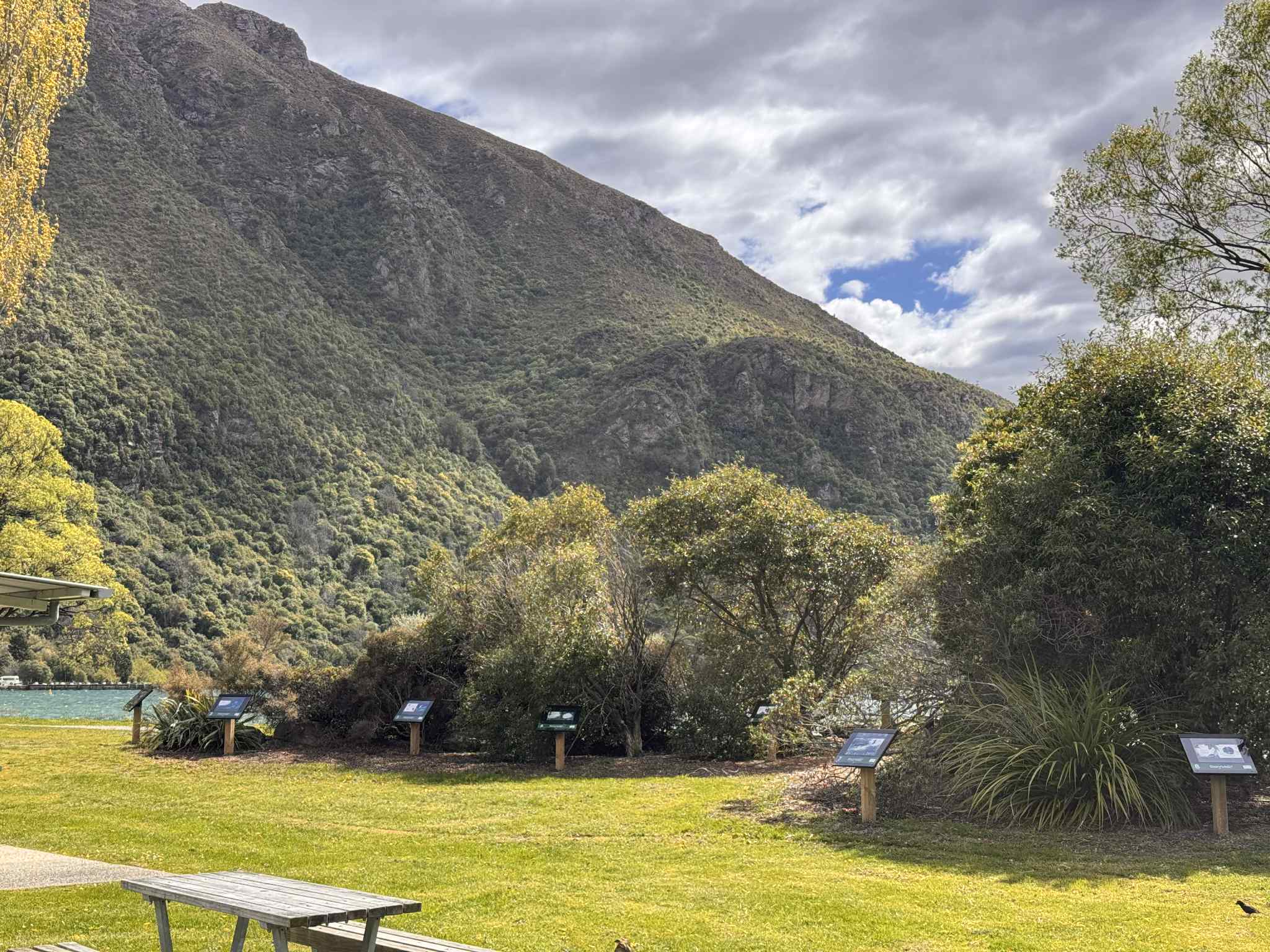 Lush green landscape with trees and story panies in a park, set against a backdrop of a large, rugged mountain under a cloudy sky.